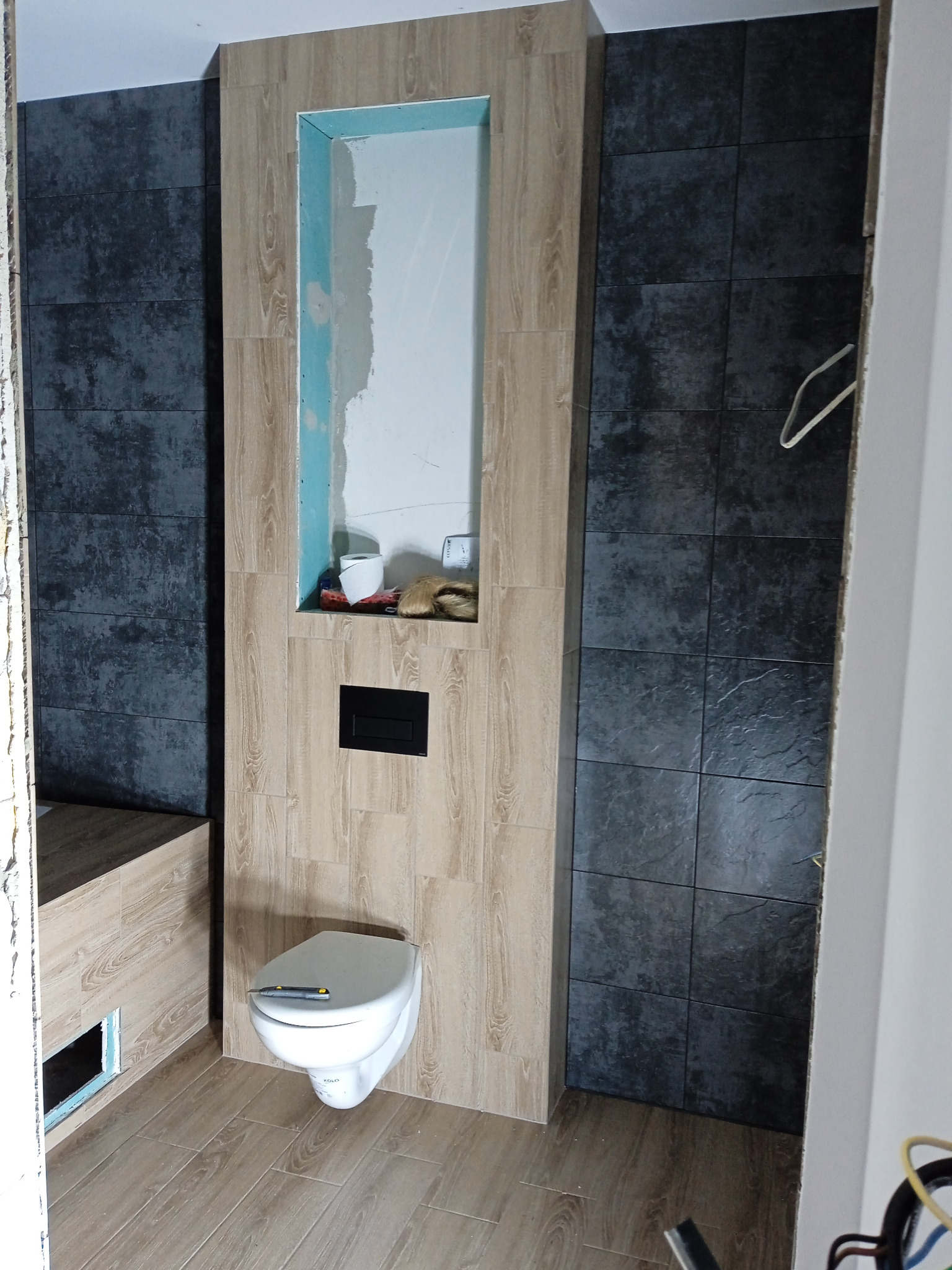 Wnętrze łazienki w trakcie prac wykończeniowych; widoczna podwieszana toaleta z nożykiem na desce, obudowa stelaża z płytek imitujących drewno z wnęką, ściany wyłożone ciemnymi płytkami.