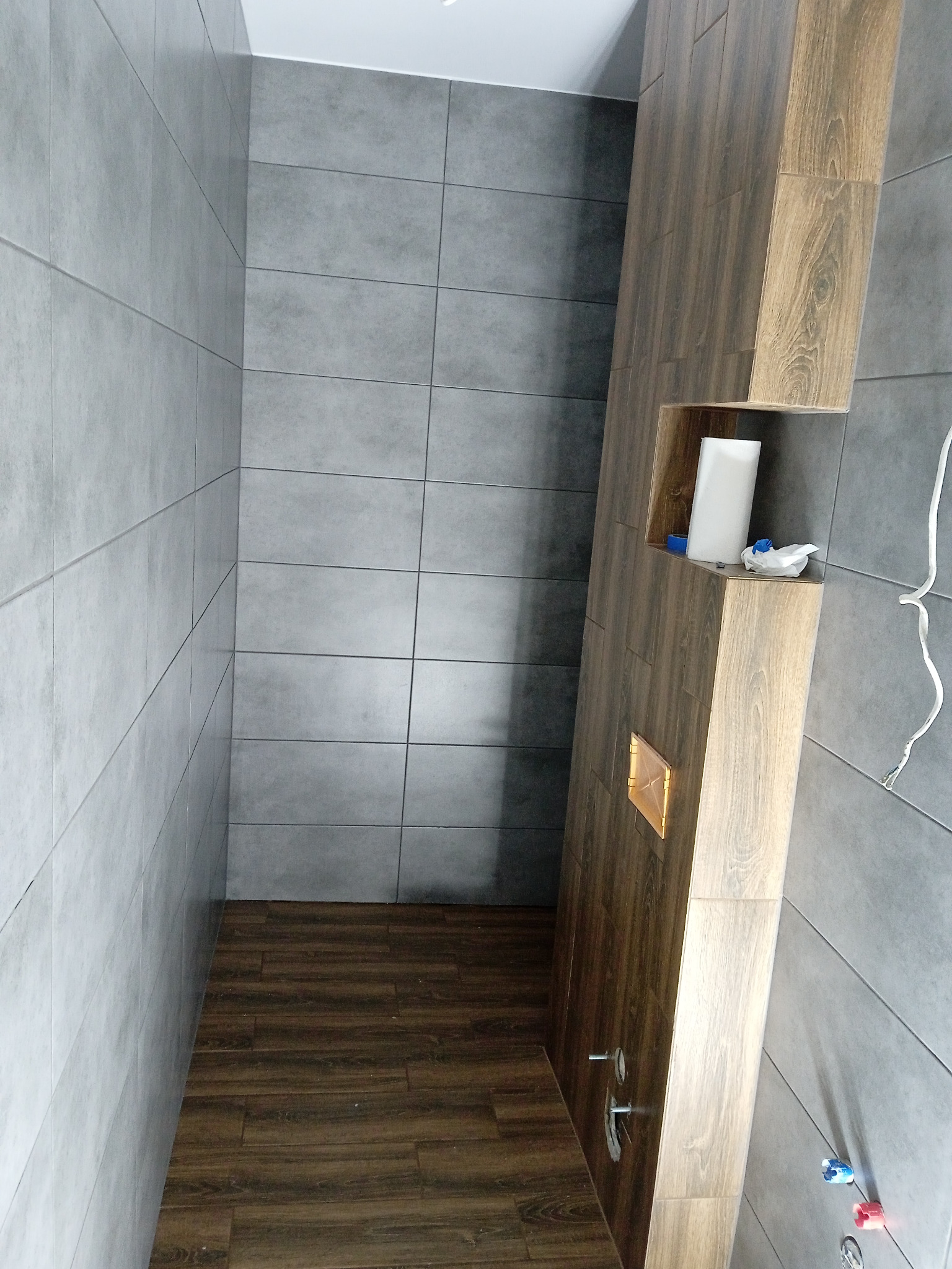 Wykończona łazienka z szarymi płytkami ściennymi i drewnopodobną zabudową z wnęką, w której stoją pojemniki. Na podłodze brązowe płytki imitujące drewno.