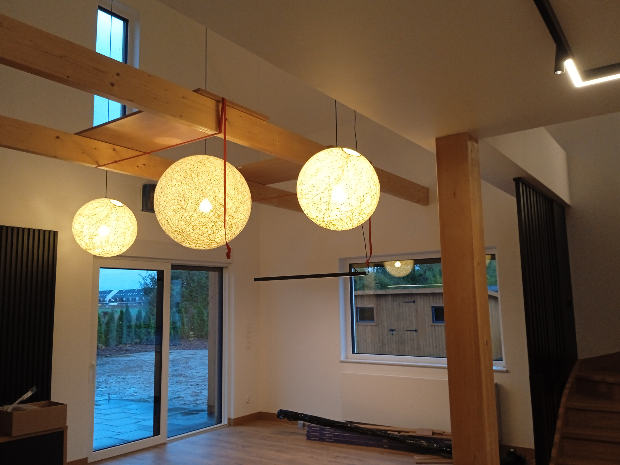 Wnętrze domu w trakcie wykańczania z drewnianymi belkami stropowymi, trzema okrągłymi lampami wiszącymi z abażurami ze sznurka i dużymi oknami przesuwnymi z widokiem na ogród. Na podłodze leżą...