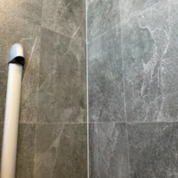 Szare, matowe płytki imitujące kamień na ścianie, pionowy odpływ liniowy z chromowaną nakładką po lewej stronie.