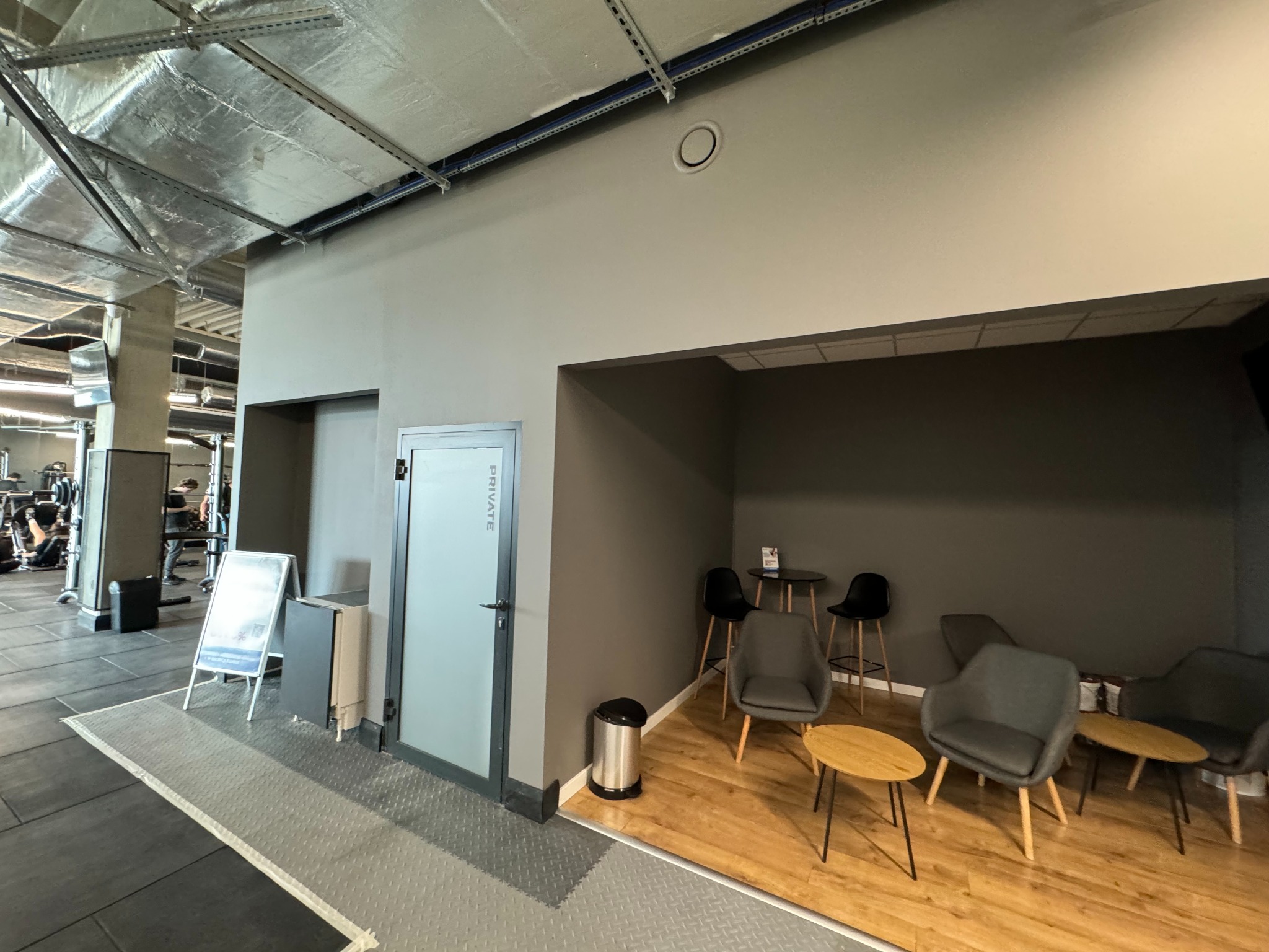 Wnętrze siłowni z szarą ścianą, aluminiowym sufitem i strefą relaksu z krzesłami i stolikami na drewnianej podłodze.