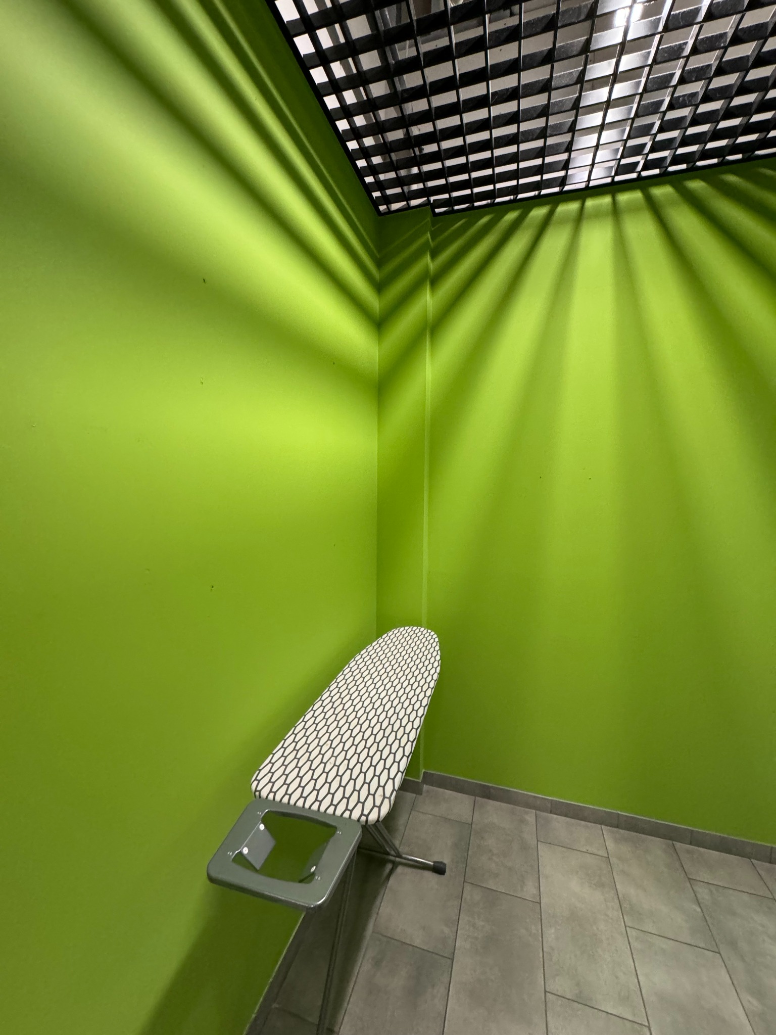 Ściana pomalowana na jaskrawy zielony kolor w rogu pokoju, z widoczną deską do prasowania i cieniem kratownicy na suficie.