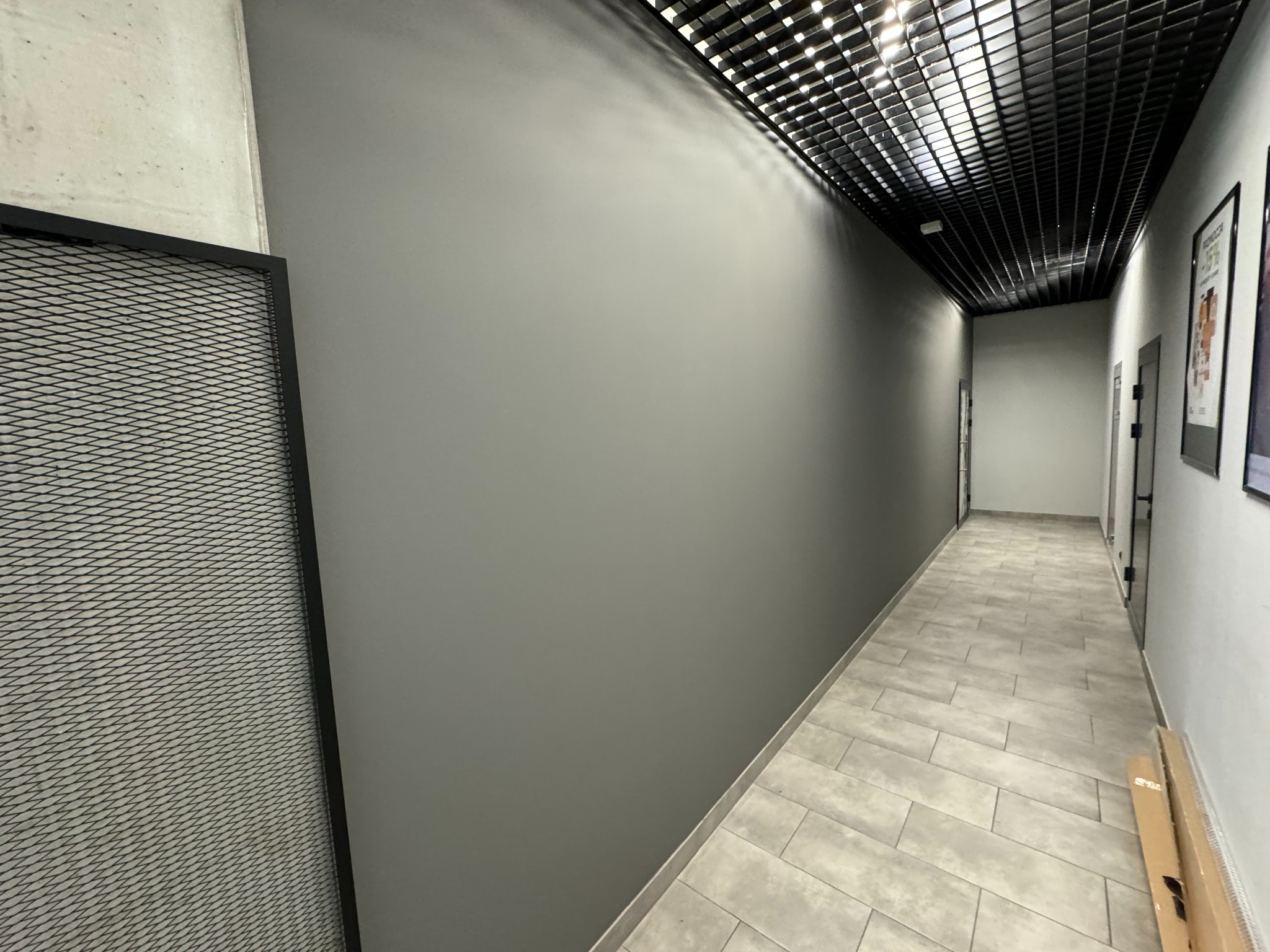 Korytarz biurowy z szarą, świeżo pomalowaną ścianą, wykończony płytkami podłogowymi w odcieniach szarości i czarnym sufitem rastrowym z oświetleniem.