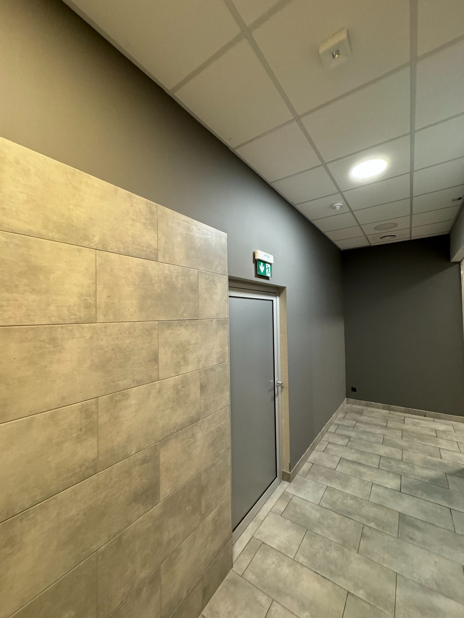 Korytarz biurowy z szarymi ścianami, beżowymi płytkami na ścianie po lewej i na podłodze, oraz szarymi drzwiami z zielonym znakiem ewakuacyjnym.
