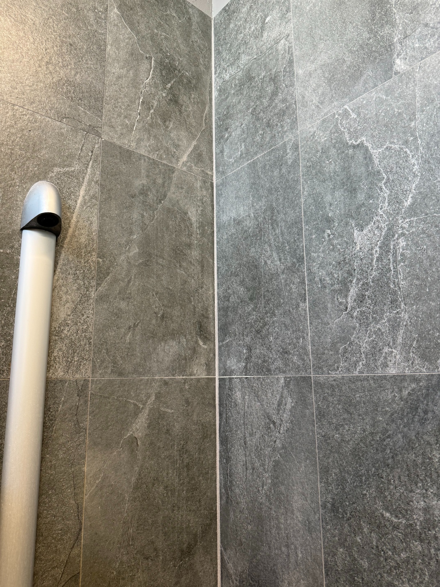 Szare, matowe płytki imitujące kamień na ścianie, pionowy odpływ liniowy z chromowaną nakładką po lewej stronie.