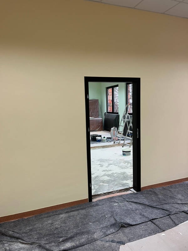 Świeżo pomalowane ściany w jasnym odcieniu kremu, widok na sąsiednie pomieszczenie przez czarne futryny drzwi, zabezpieczona podłoga w pomieszczeniu obok, drabina i akcesoria malarskie w tle.