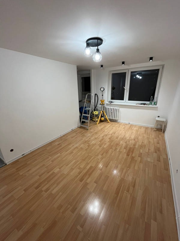 Wnętrze pokoju po świeżym pomalowaniu ścian na biało, widoczny parkiet, drabina, odkurzacz przemysłowy i lampa sufitowa z trzema kloszami.