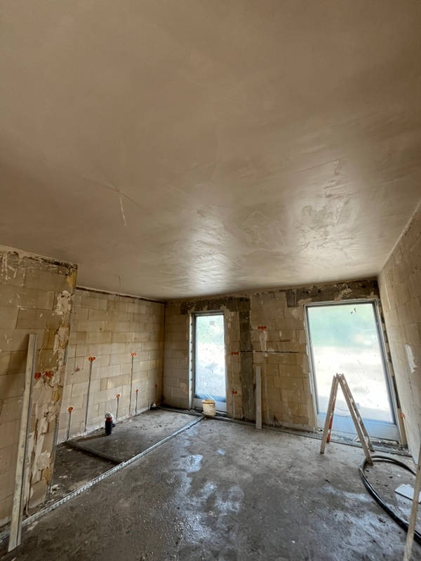 Pokój w trakcie prac wykończeniowych: ściany z bloków, częściowo pokryte tynkiem, widoczne instalacje elektryczne, dwa okna zabezpieczone folią, na podłodze resztki zaprawy i listwy przypodłogowe.