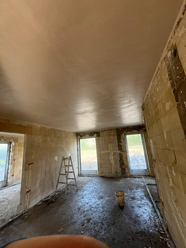 Surowe wnętrze w trakcie prac wykończeniowych: ściany z betonu komórkowego po nałożeniu warstwy tynku, widoczna drabina i wiadro na mokrej podłodze, okna bez parapetów.