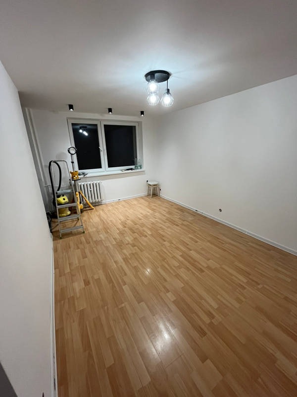 Pokój po odświeżeniu ścian na biało, widoczna drabina, grzejnik, drewniana podłoga i nowoczesna lampa sufitowa z trzema żarówkami.