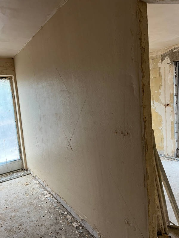 Świeżo nałożona, jasna warstwa gładzi na ścianie w trakcie remontu, widoczne ślady po szpachli, surowy beton na podłodze, okno w tle.