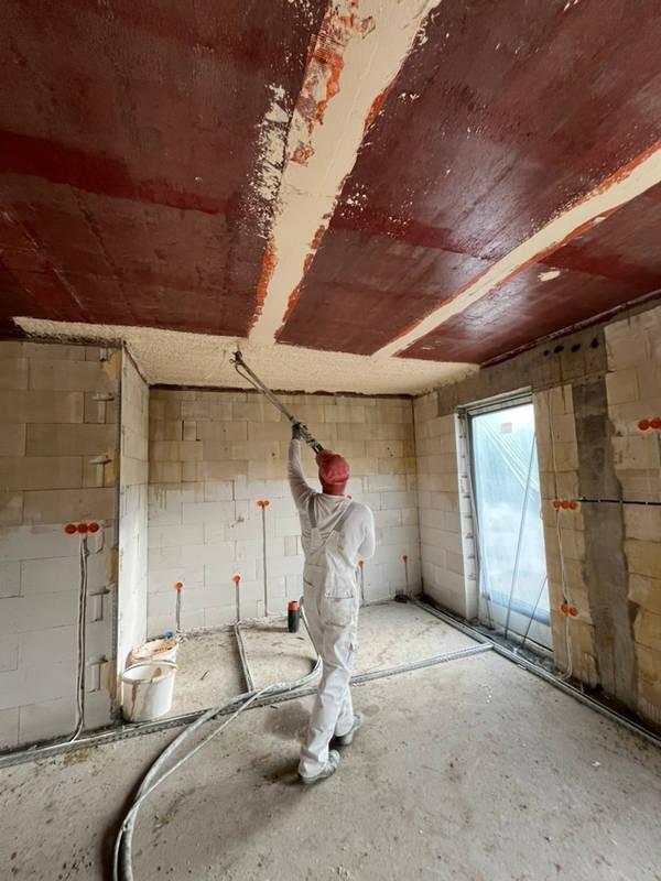 Tynkarz w kombinezonie i czerwonej czapce nanosi tynk maszynowy na sufit w surowym wnętrzu z instalacją elektryczną w ścianach.