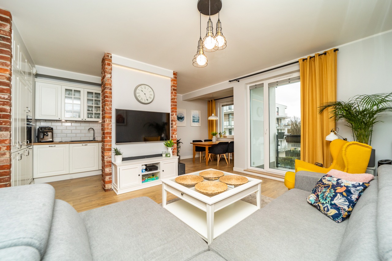 Jasne, przestronne wnętrze mieszkania z ceglanymi elementami, białymi meblami kuchennymi, szarą sofą z kolorową poduszką, żółtym fotelem i zasłonami, oraz stołem jadalnym w tle.