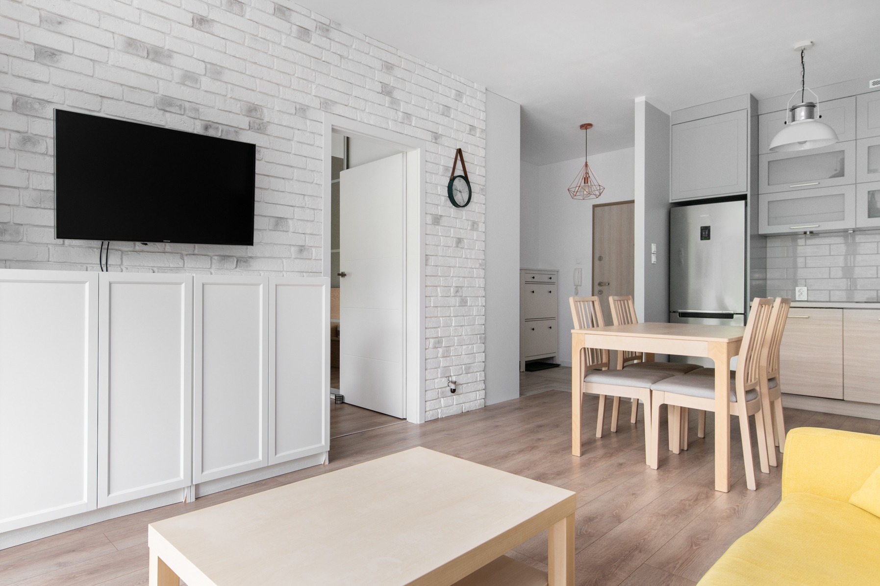Jasne, przestronne wnętrze mieszkania z ceglaną ścianą, telewizorem, białymi meblami, drewnianym stołem z krzesłami oraz fragmentem żółtej sofy.