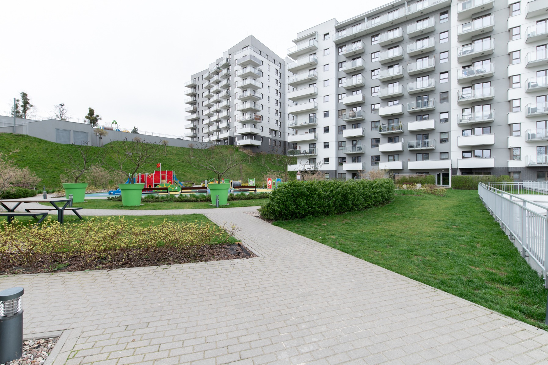 Nowoczesne osiedle mieszkaniowe z placem zabaw, widok z brukowanej ścieżki, zieleń trawników i krzewów, duże donice w kolorze zielonym.