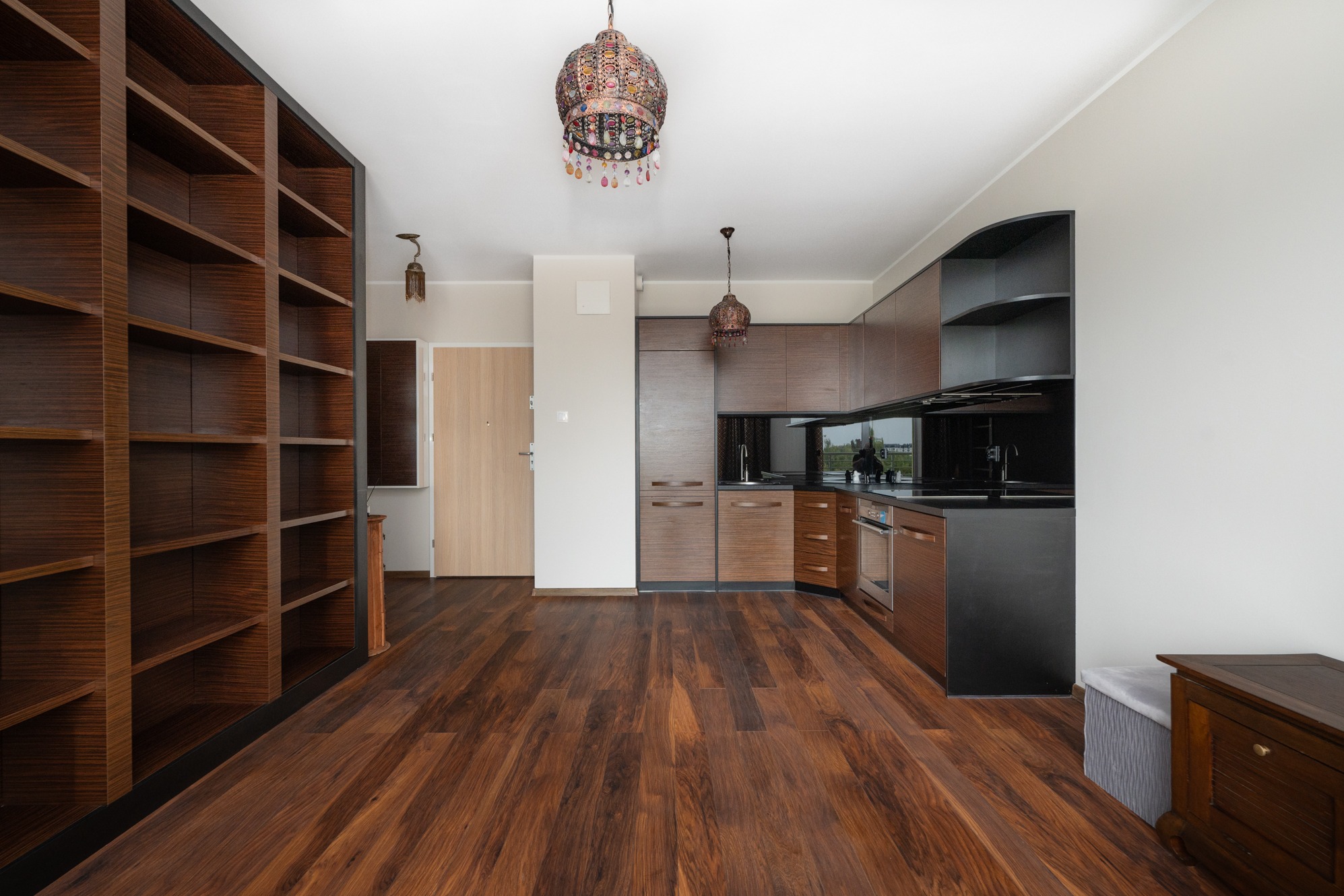 Nowoczesne wnętrze mieszkania z drewnianą podłogą, zabudową kuchenną w odcieniach brązu i czerni, oraz wysokim regałem na książki. Dwa ozdobne żyrandole wiszą pod sufitem. Widok na okno zza...