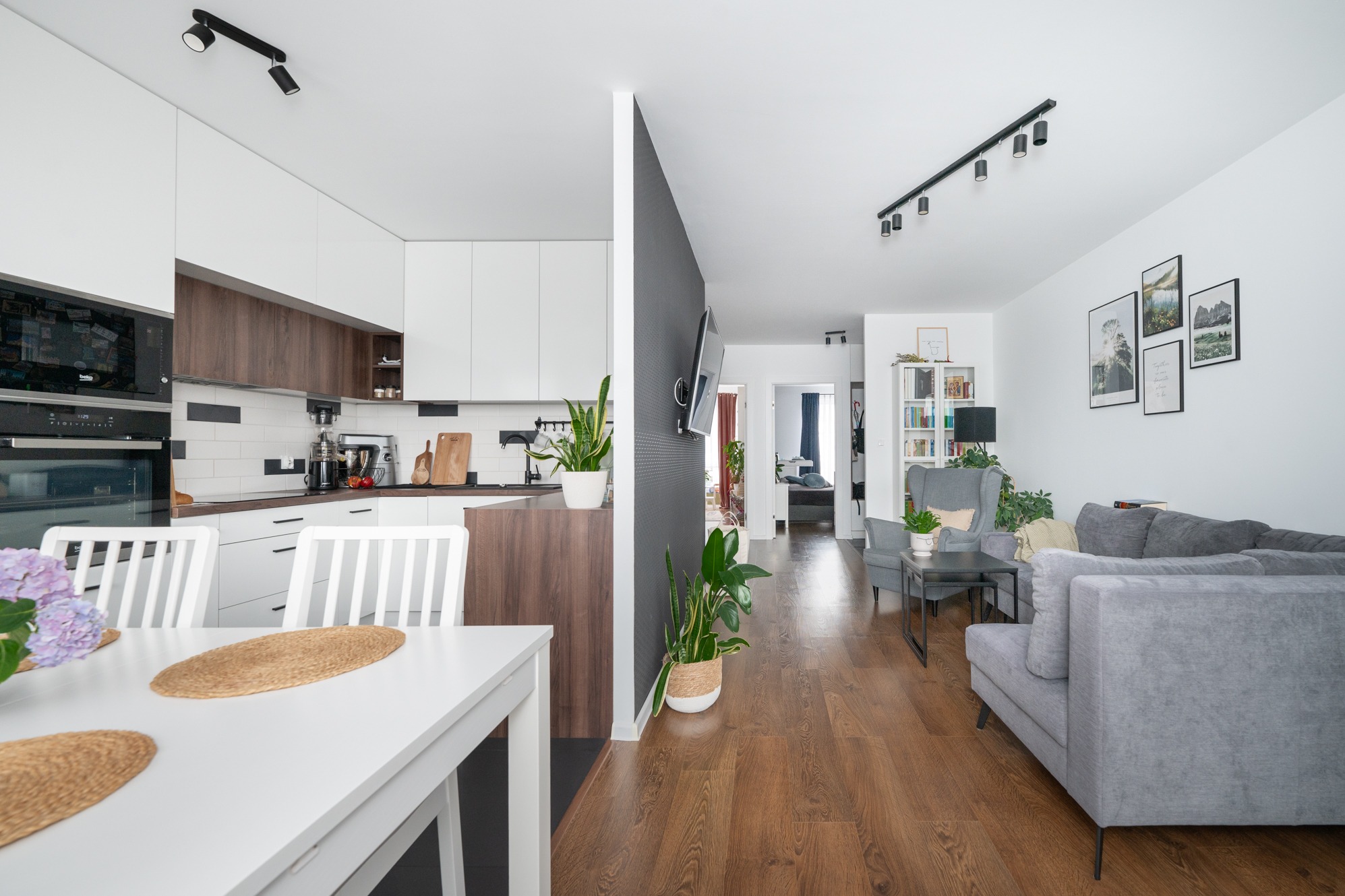 Jasne, przestronne wnętrze mieszkania z otwartą kuchnią, białymi szafkami i drewnianymi akcentami, widok na salon z szarą sofą i fotelami, drewniana podłoga, nowoczesne oświetlenie punktowe.