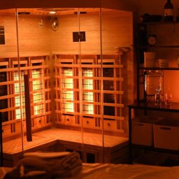 Gabinet masażu zdrowia i relaksu GMRonczek - Wnętrze sauny infrared z drewnianymi panelami i szklanymi drzwiami, oświetlone ciepłym światłem, obok regał z dekoracjami i naczyniami.