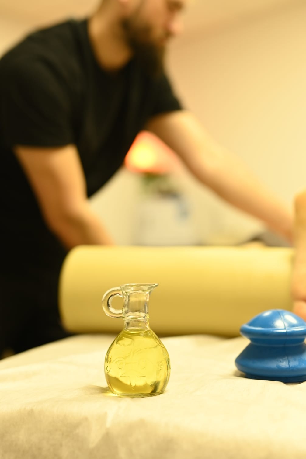 Olej do masażu w szklanej karafce z uchwytem, obok niebieskie bańki chińskie, w tle osoba wykonująca zabieg na leżance z wałkiem pod kolanami.