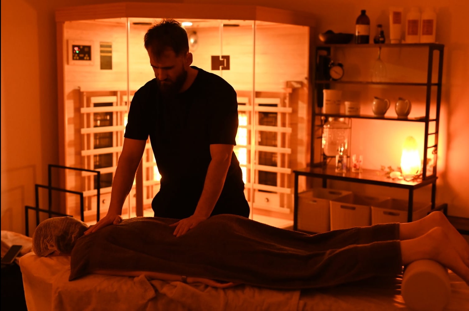 Mężczyzna wykonuje masaż pleców pacjentki leżącej na stole w gabinecie z sauną infrared w tle i półkami z kosmetykami.