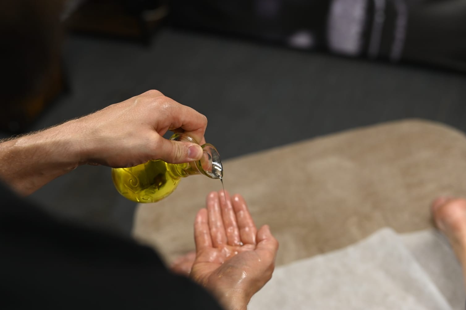 Olej wylewany z małej szklanej karafki na dłoń osoby leżącej na leżance pokrytej beżowym i białym materiałem.