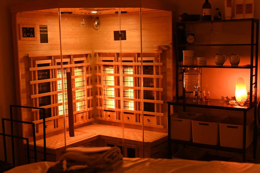 Wnętrze sauny infrared z drewnianymi panelami i szklanymi drzwiami, oświetlone ciepłym światłem, obok regał z dekoracjami i naczyniami.