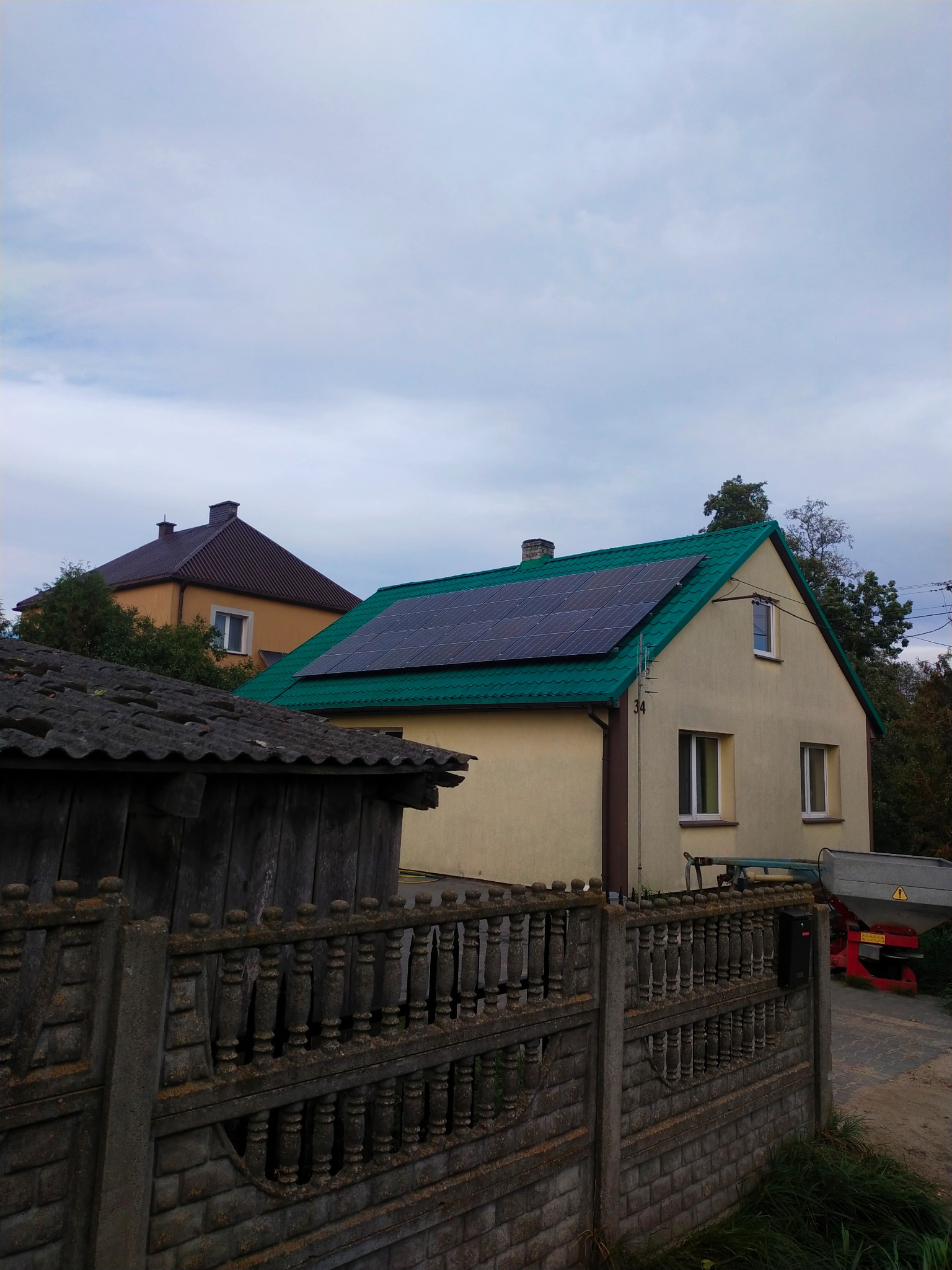 Dom jednorodzinny z zielonym dachem, na którym zamontowano panele słoneczne; widok zza betonowego ogrodzenia.