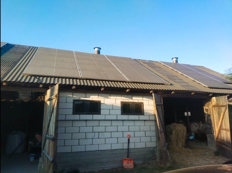 Instalacja paneli słonecznych na dachu starego budynku gospodarczego z białej cegły, widoczna szopa z otwartymi drzwiami, wewnątrz siano, na dachu kominy wentylacyjne.