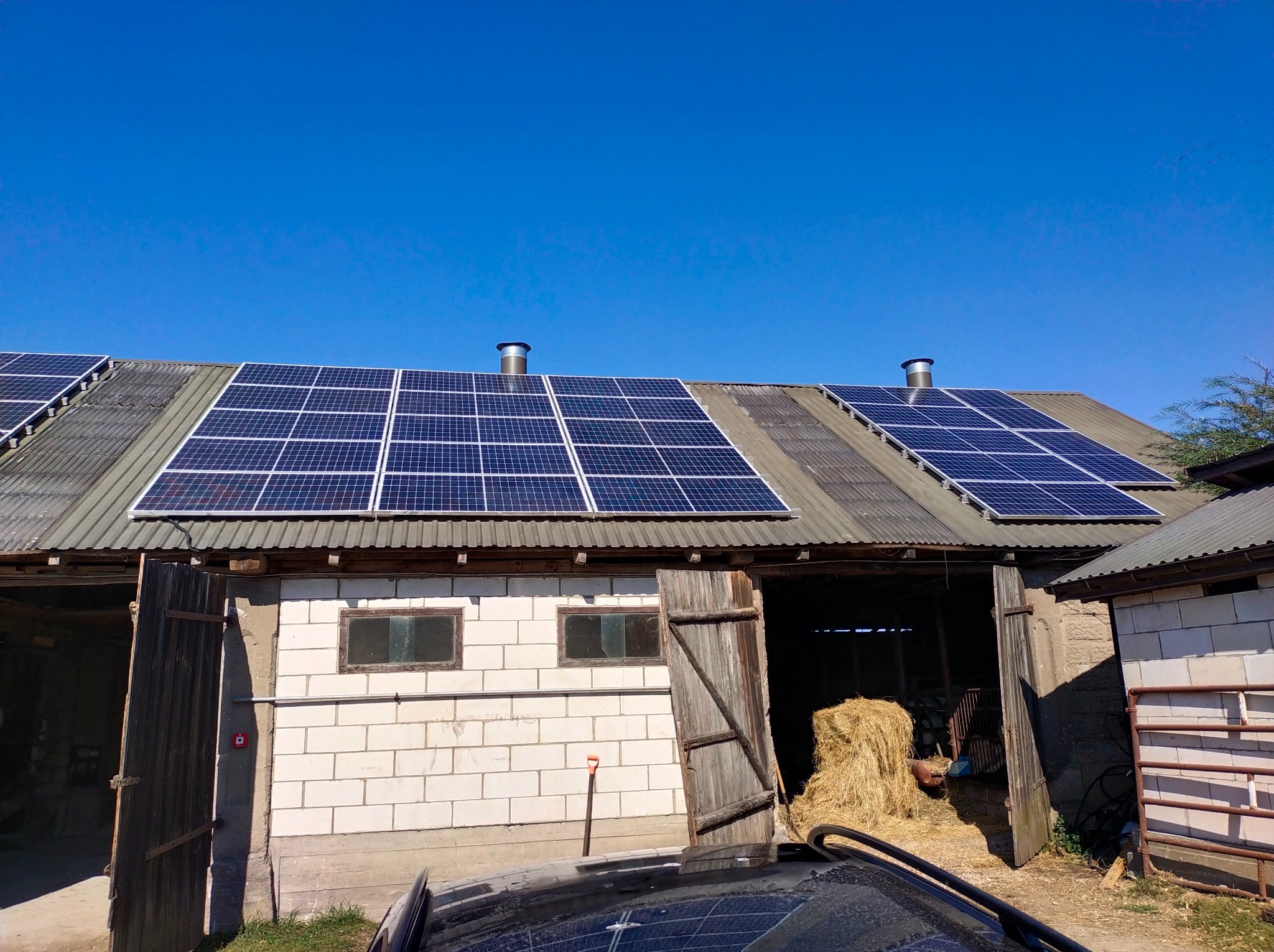 Panele słoneczne na dachu starego budynku gospodarczego z otwartymi drewnianymi wrotami, wewnątrz widoczne bele siana, na dachu dwa kominki wentylacyjne, błękitne niebo w tle.