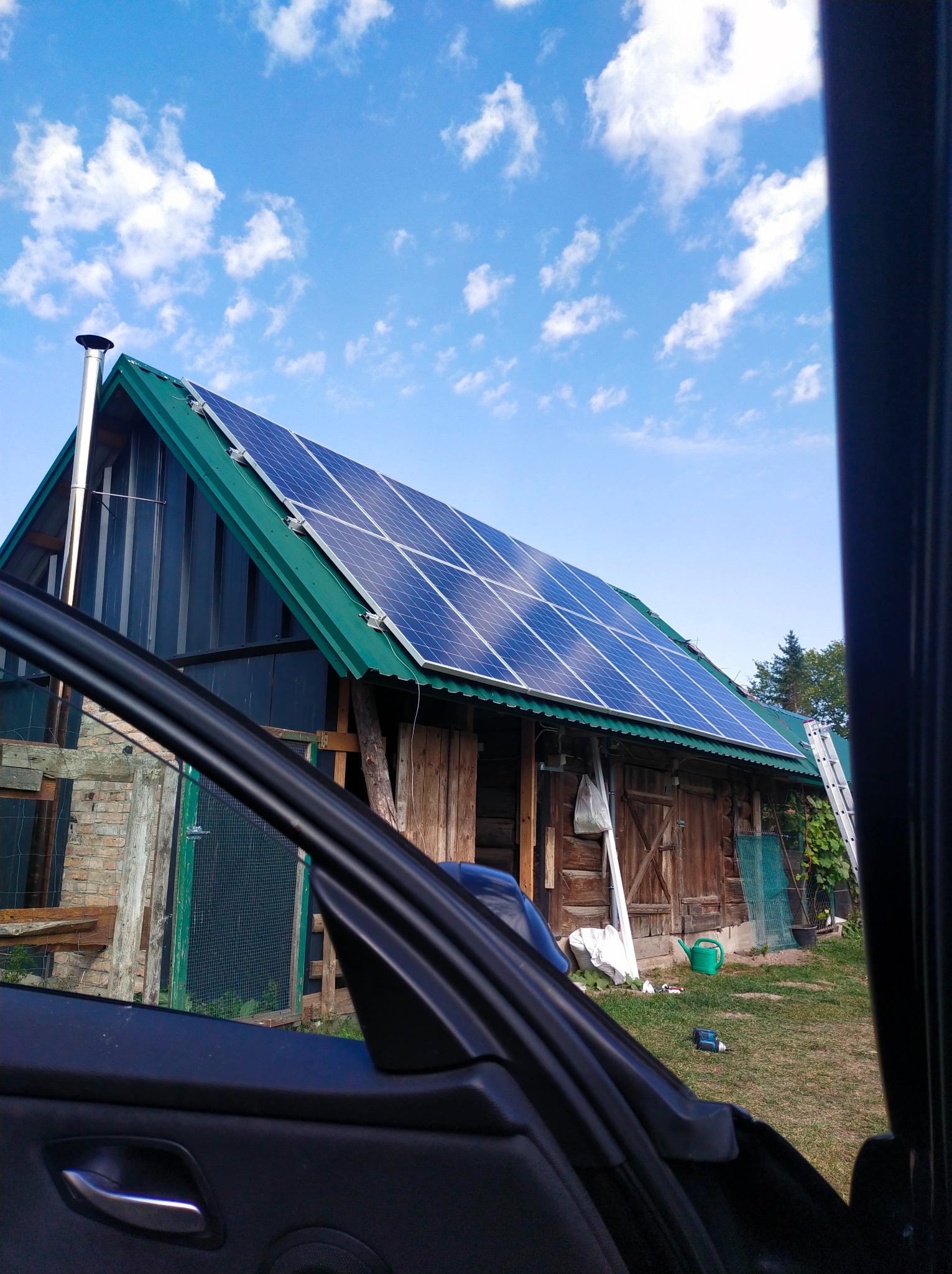 Widok z wnętrza auta na drewniany budynek z zielonym dachem, na którym zamontowane są panele słoneczne, z kominem i drabiną opartą o ścianę, na tle błękitnego nieba z chmurami.