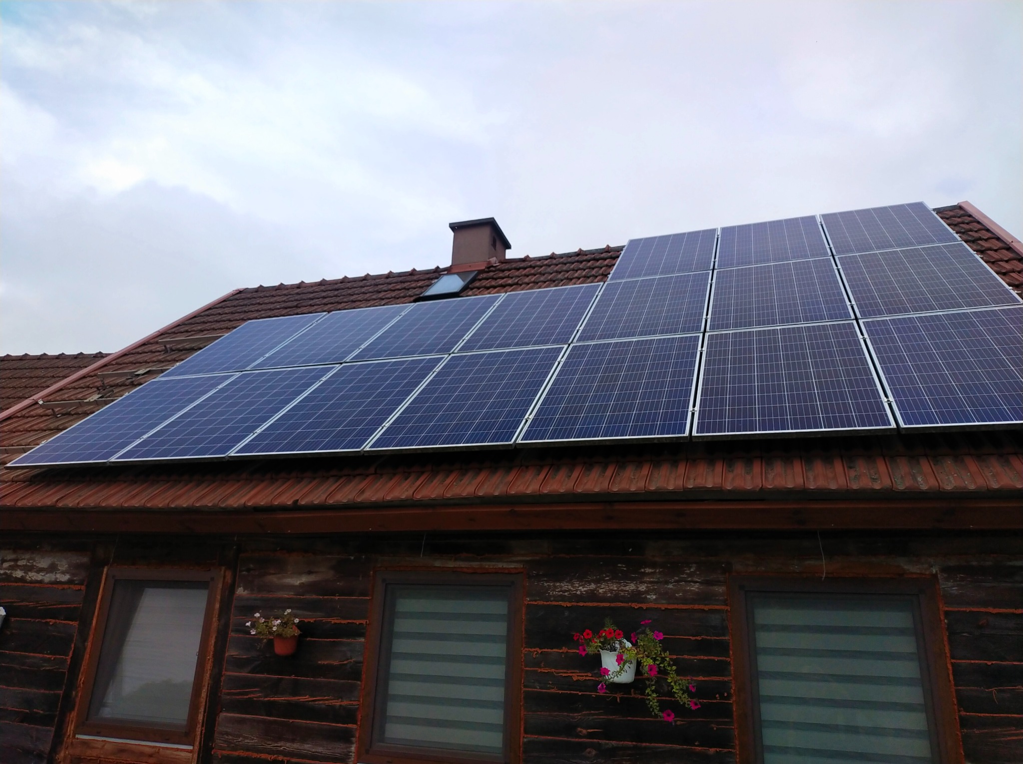 Panele fotowoltaiczne na dachu drewnianego domu z czerwonej dachówki, widoczne okna z kwiatami w doniczkach.