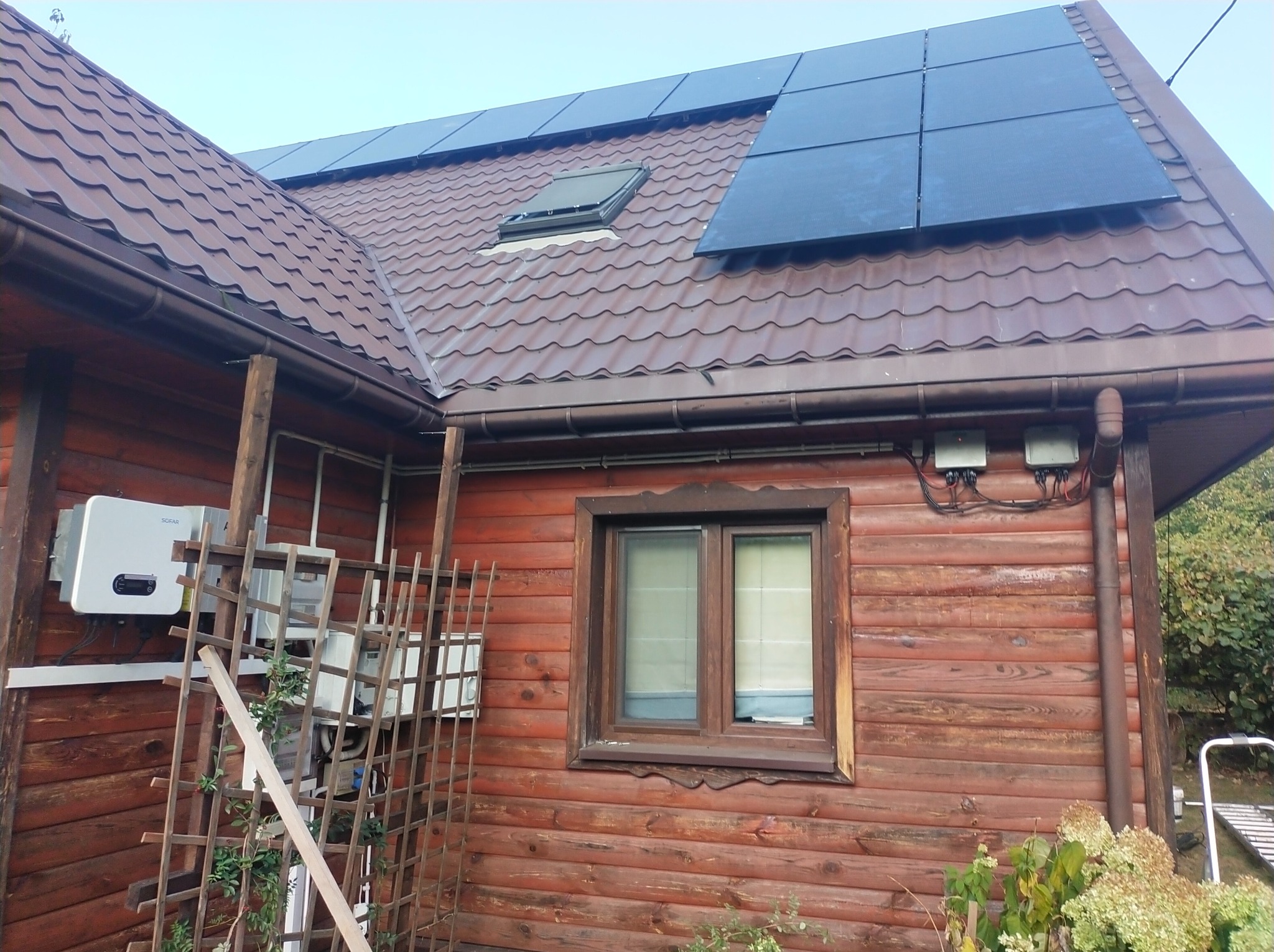 Dom z brązową dachówką, oknem dachowym i panelami słonecznymi; widoczne również dwa inwertery na drewnianej elewacji oraz elementy instalacji elektrycznej na zewnątrz.