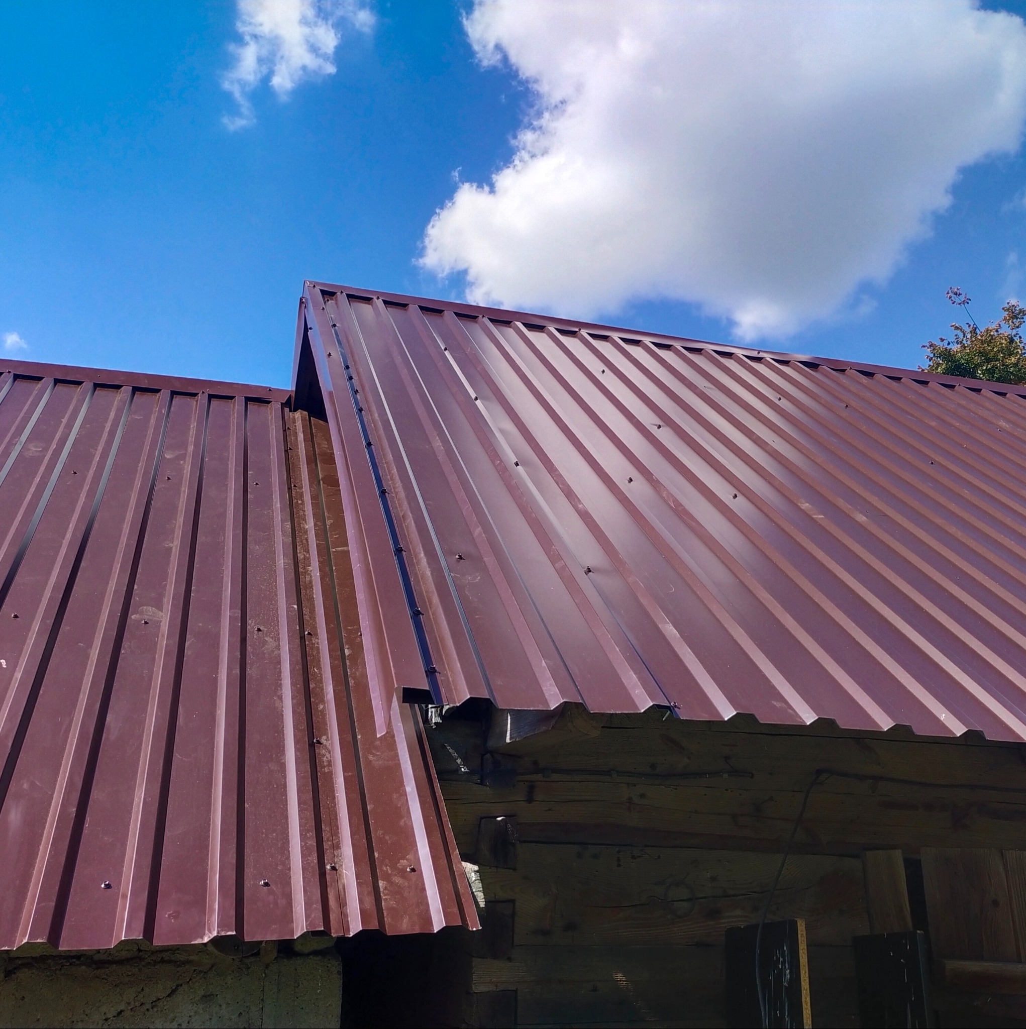 Ciemnoczerwone, trapezowe blachy dachowe na drewnianej konstrukcji, widok z dołu na tle błękitnego nieba z chmurami.