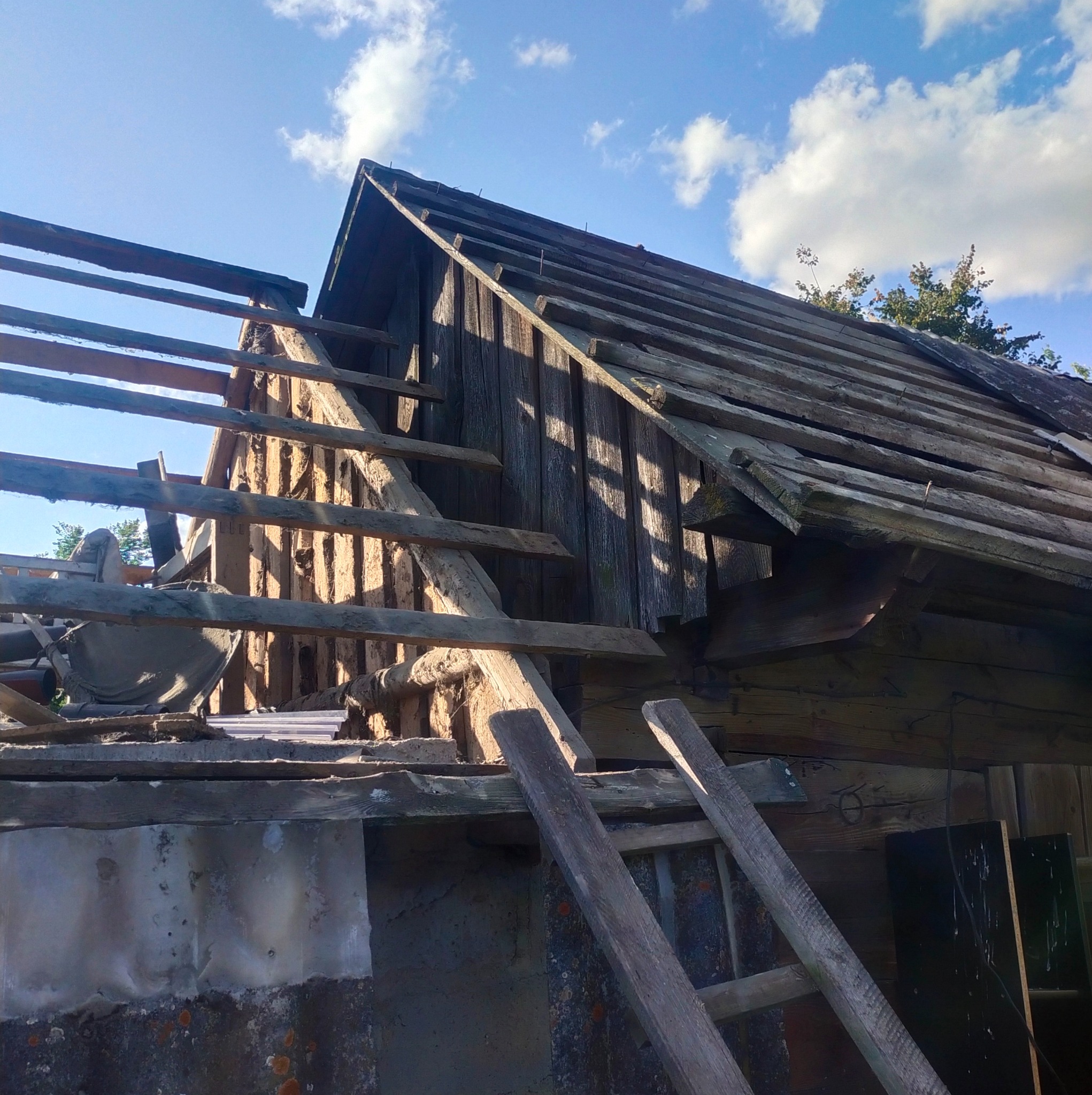 Demontaż starego, drewnianego dachu z widoczną konstrukcją więźby i fragmentami eternitu na ścianie, z niebem i chmurami w tle.