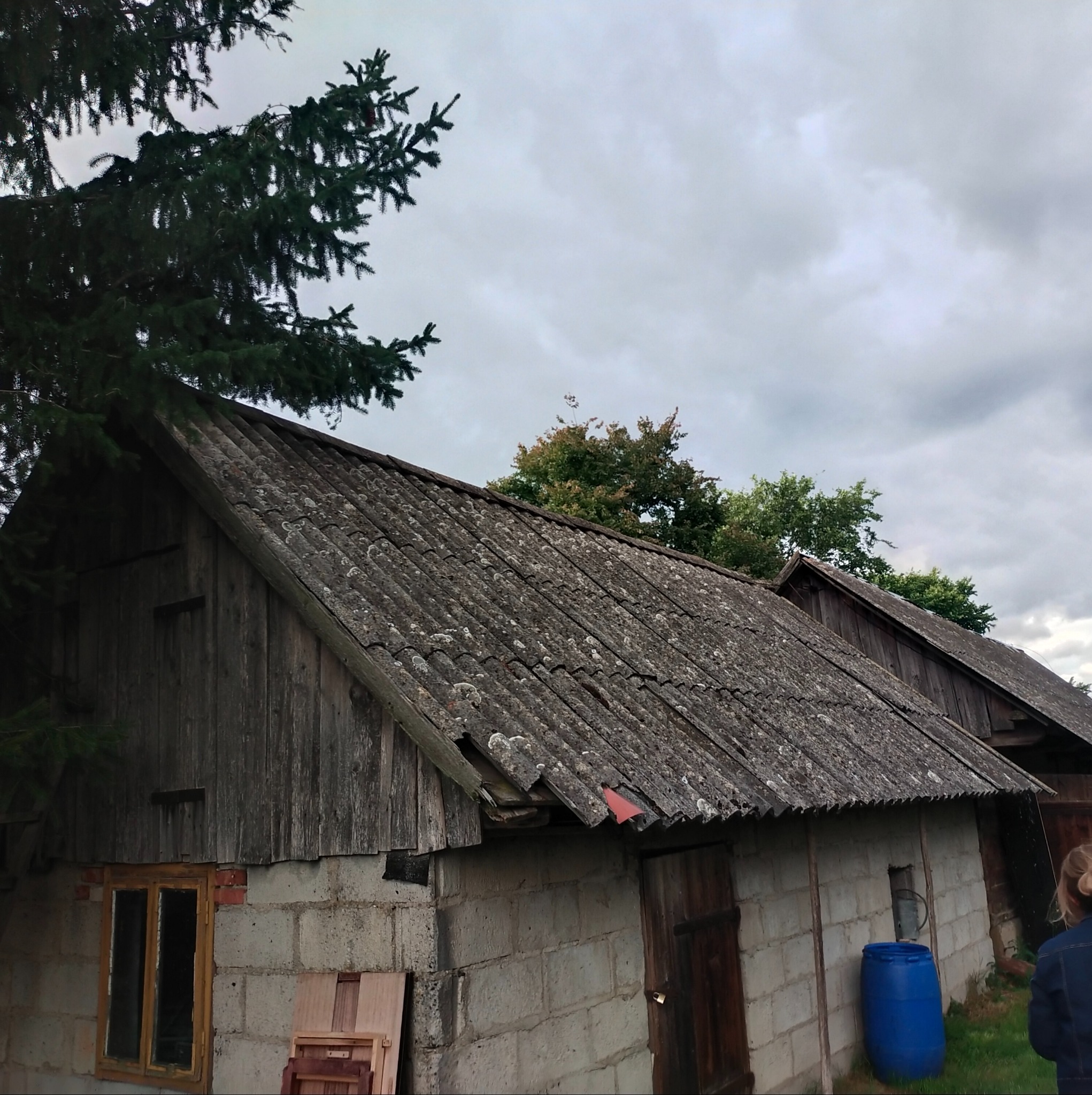 Stary budynek gospodarczy z szarą, zniszczoną dachówką eternitową pokrytą mchem, widoczne ślady uszkodzeń i brakujących elementów, ściany z bloków betonowych, drewniane elementy konstrukcyjne...