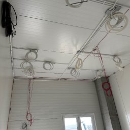 INSTALELEKTRO24 PROSTA SPÓŁKA AKCYJNA - Instalacja elektryczna w hali: widok na sufit z zamocowanymi rurami PCV, przewodami elektrycznymi w kolorach białym i czerwonym, oraz czarną wiązką kabli przy ścianie. Widoczny betonowy filar...