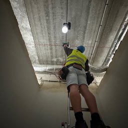 INSTALELEKTRO24 PROSTA SPÓŁKA AKCYJNA - Elektryk w kamizelce odblaskowej i niebieskim kasku na drabinie, montuje oświetlenie w surowym wnętrzu, widoczne przewody i rury wentylacyjne pod betonowym stropem.
