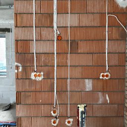 INSTALELEKTRO24 PROSTA SPÓŁKA AKCYJNA - Montaż przewodów elektrycznych w nowym budynku z cegły, widoczne puszki instalacyjne i kable prowadzone w bruzdach, surowy stan wykończenia.