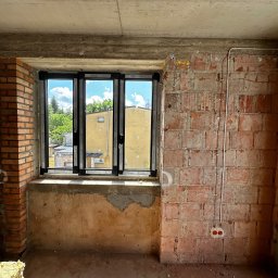 INSTALELEKTRO24 PROSTA SPÓŁKA AKCYJNA - Surowy stan wnętrza z oknem i widoczną instalacją elektryczną w peszlach na ceglanej ścianie, gniazdko elektryczne w puszce.