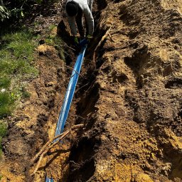 INSTALELEKTRO24 PROSTA SPÓŁKA AKCYJNA - Pracownik w szarej bluzie układa niebieski przewód w wykopie ziemnym, widoczne korzenie i roślinność.