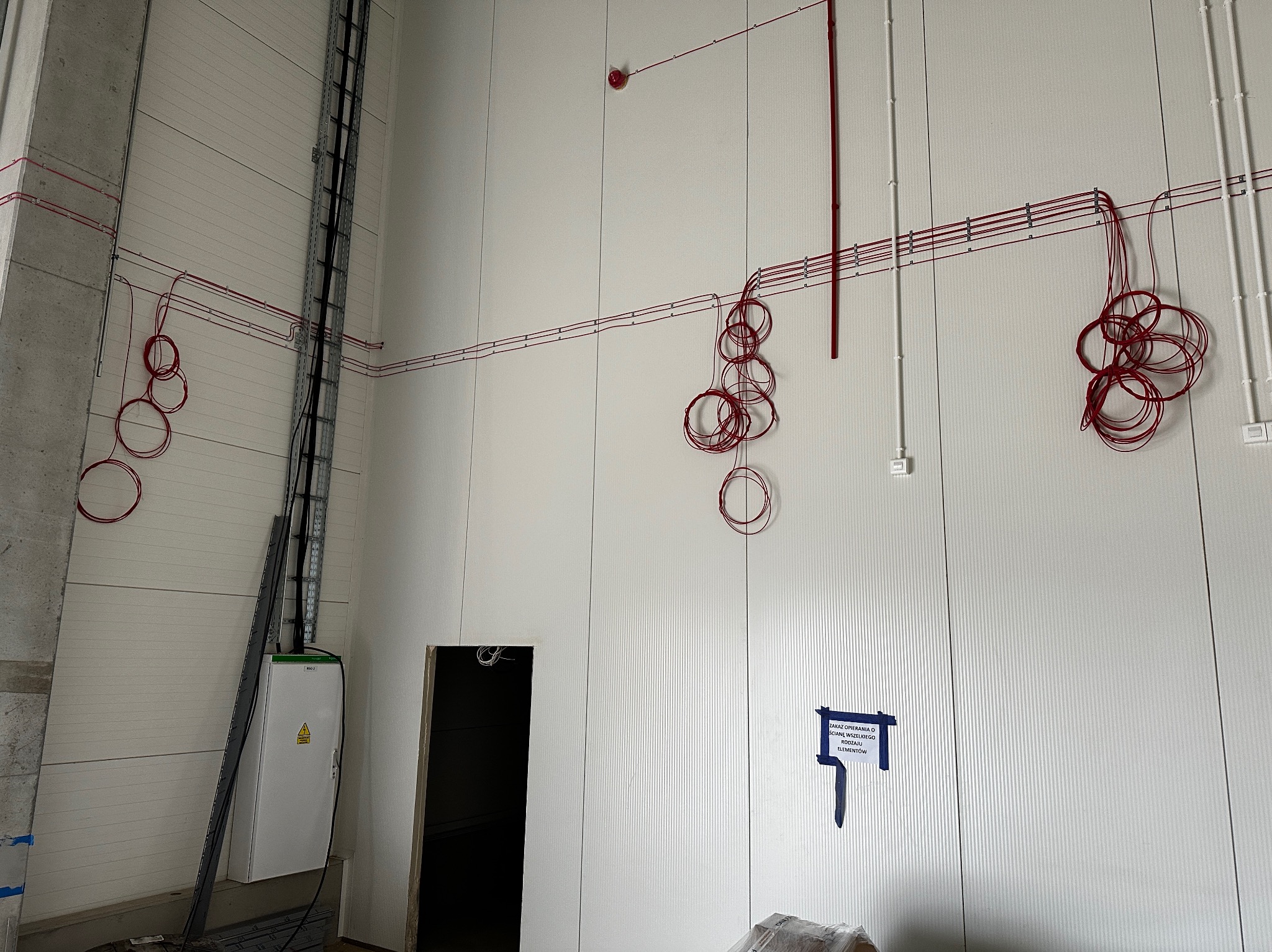 Wnętrze hali produkcyjnej w trakcie instalacji elektrycznej: zwisające kable w pętlach, pionowy kanał kablowy, skrzynka elektryczna z ostrzeżeniem, otwór drzwiowy i tabliczka informacyjna na białej...