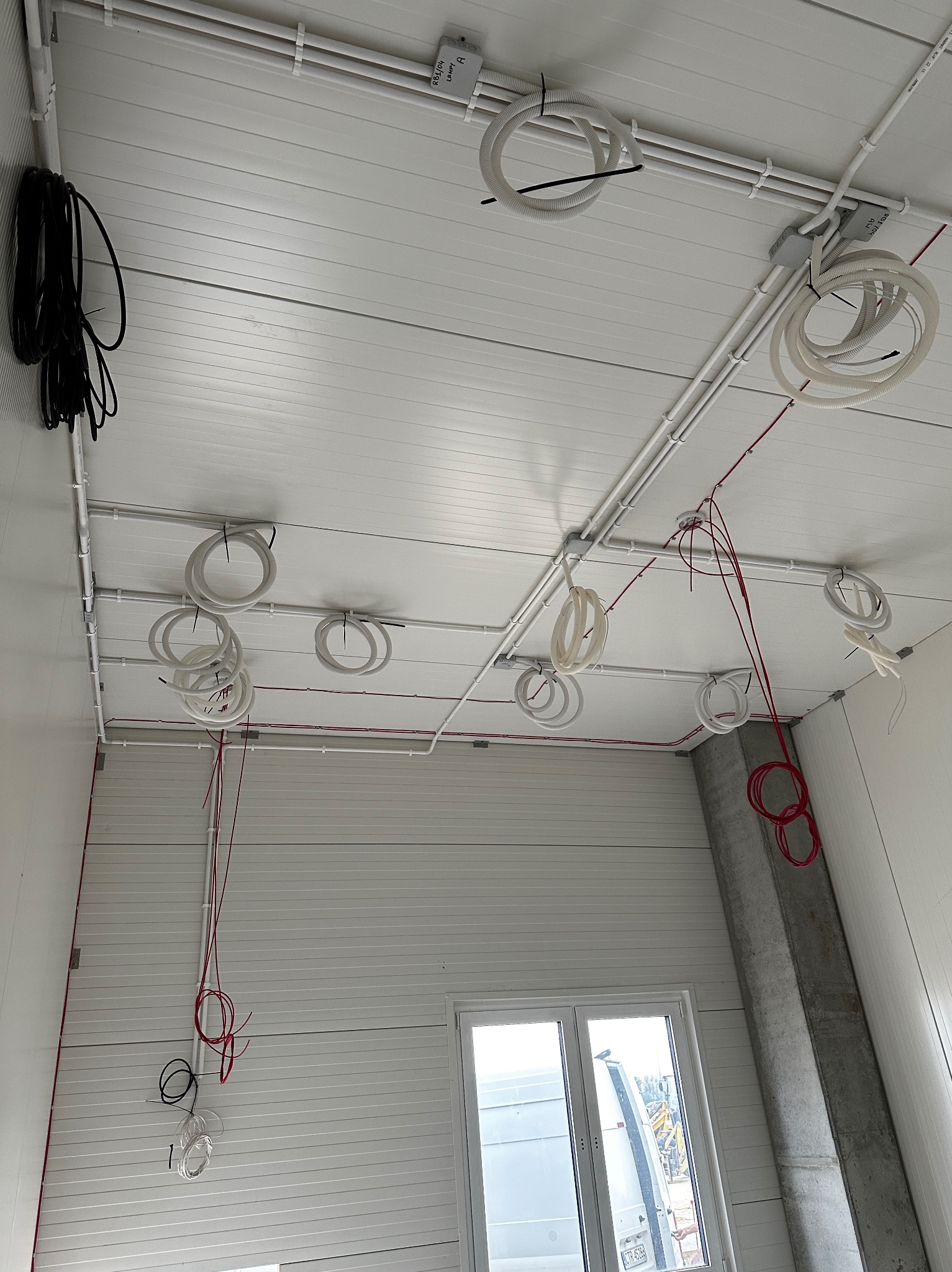 Instalacja elektryczna w hali: widok na sufit z zamocowanymi rurami PCV, przewodami elektrycznymi w kolorach białym i czerwonym, oraz czarną wiązką kabli przy ścianie. Widoczny betonowy filar...