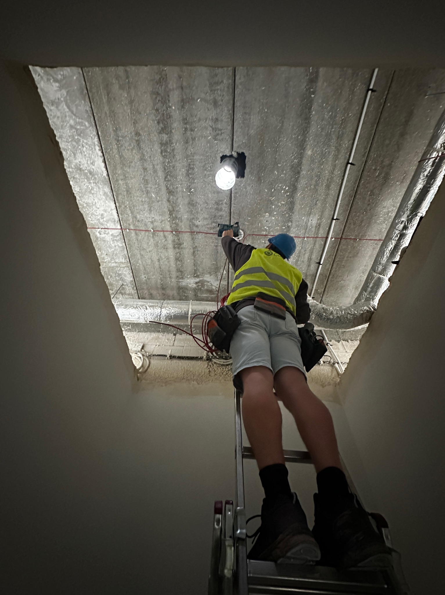 Elektryk w kamizelce odblaskowej i niebieskim kasku na drabinie, montuje oświetlenie w surowym wnętrzu, widoczne przewody i rury wentylacyjne pod betonowym stropem.