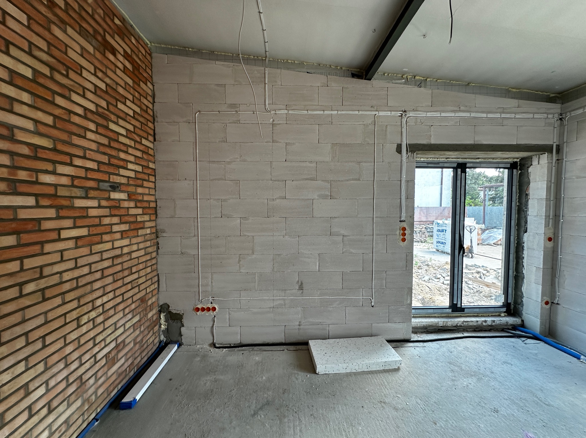 Surowy stan wnętrza z instalacją elektryczną: widoczne kable i puszki na ścianie z betonu komórkowego, fragment ściany z cegły klinkierowej, w tle okno z widokiem na zewnątrz.