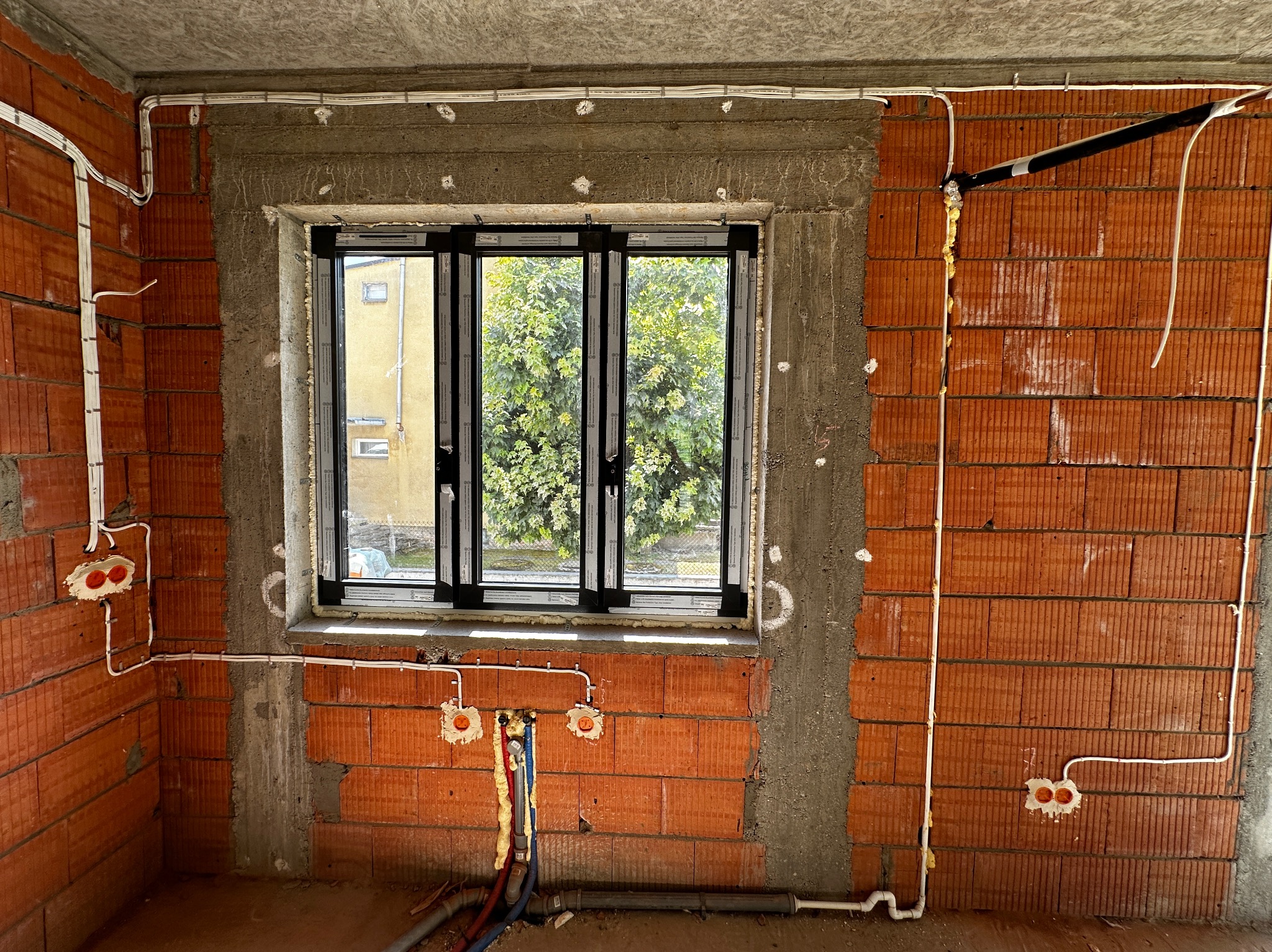 Surowy stan wnętrza z oknem: widoczne cegły, beton, rozprowadzone przewody elektryczne w białych rurkach oraz rury instalacji wodnej w podłodze.