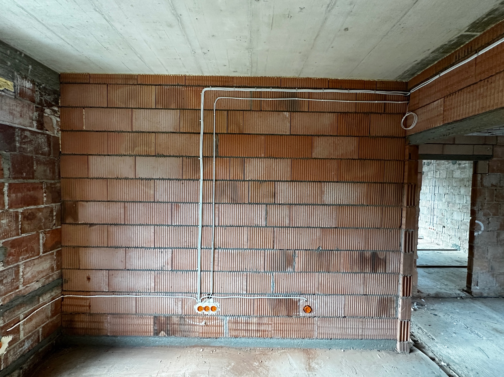 Surowy mur z pustaków ceramicznych z poprowadzonymi przewodami elektrycznymi w białych peszlach i puszkami instalacyjnymi z pomarańczowymi zaślepkami, widok na wnętrze w trakcie budowy.