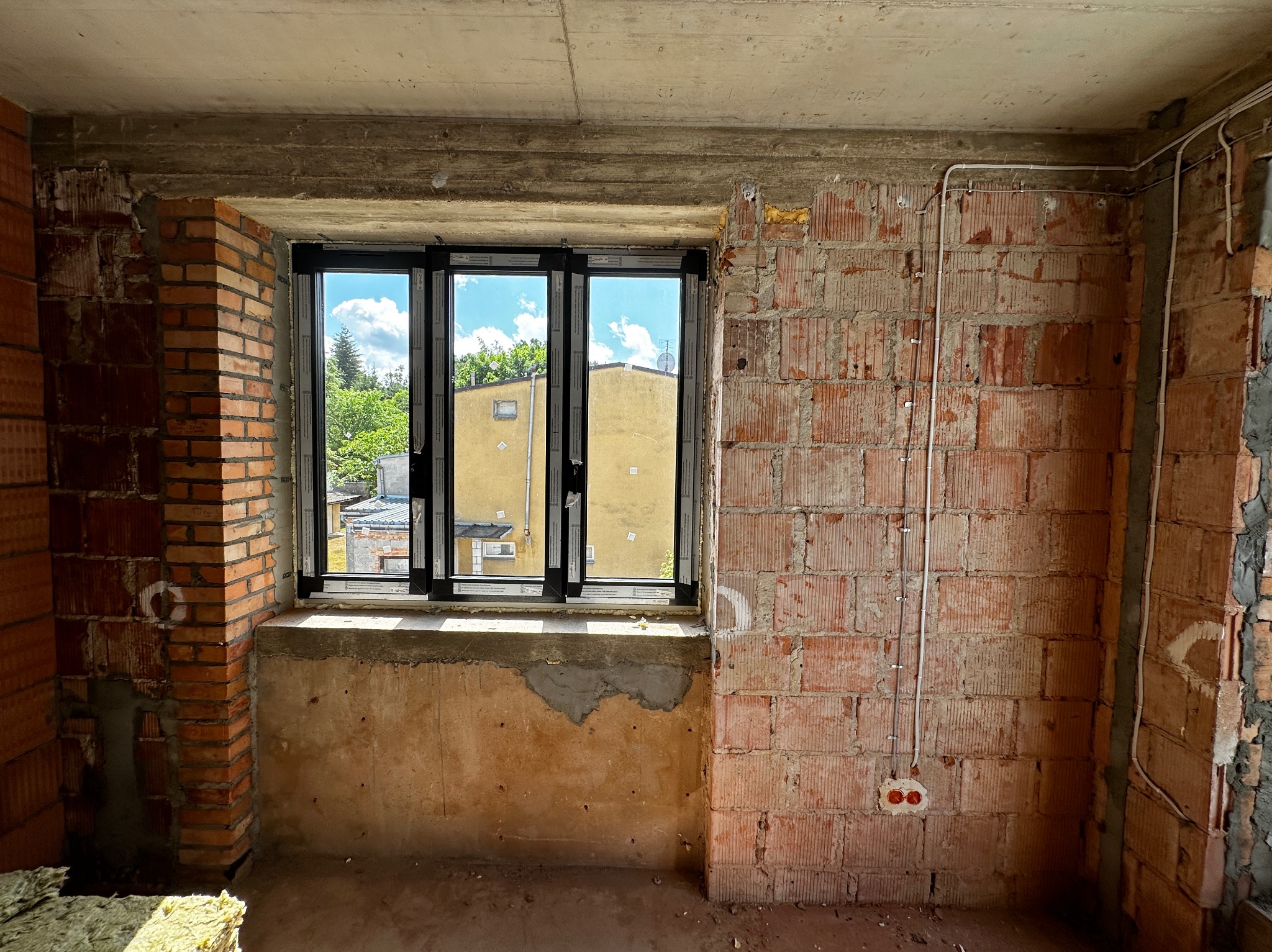 Surowy stan wnętrza z oknem i widoczną instalacją elektryczną w peszlach na ceglanej ścianie, gniazdko elektryczne w puszce.