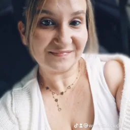 Portret kobiety w Słupsku, uśmiechniętej, z blond włosami i złotym naszyjnikiem z serduszkami, ubrana w białą koszulkę i sweter.