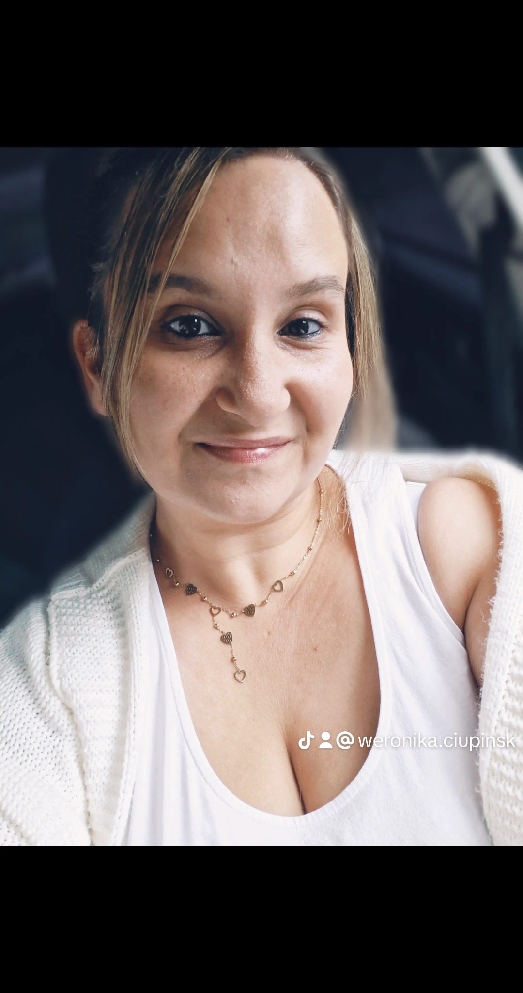 Portret kobiety w Słupsku, uśmiechniętej, z blond włosami i złotym naszyjnikiem z serduszkami, ubrana w białą koszulkę i sweter.