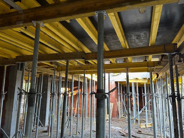 Szare, metalowe stemple budowlane podtrzymujące żółte, drewniane szalunki stropowe w trakcie budowy budynku z czerwonej cegły.